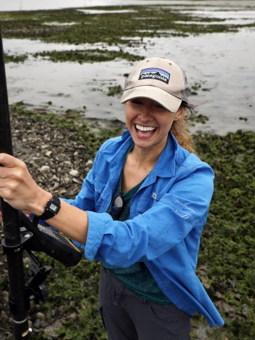Woman with hat on smiling and holding a piece of scientific equipment. Field of oysters in the background.