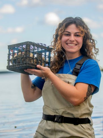 Smiling white woman with brown, curly hair holding up cage of oysters. Water in background.